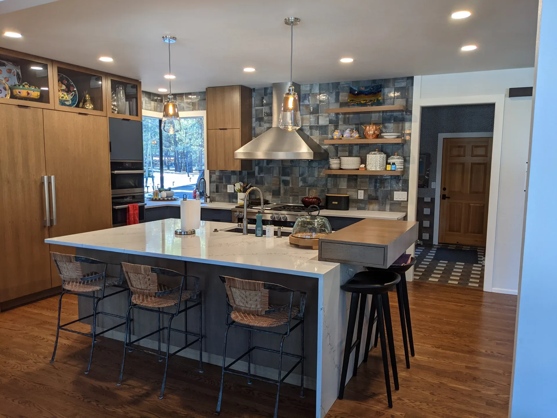 Kitchen with a white marble-look waterfall island, woven-back barstools, a stainless range hood, blue-grey tile backsplash, and light wood cabinetry